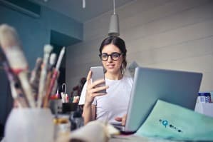 Young woman wearing glasses using a smartphone and laptop in a creative studio with paintbrushes in the foreground.