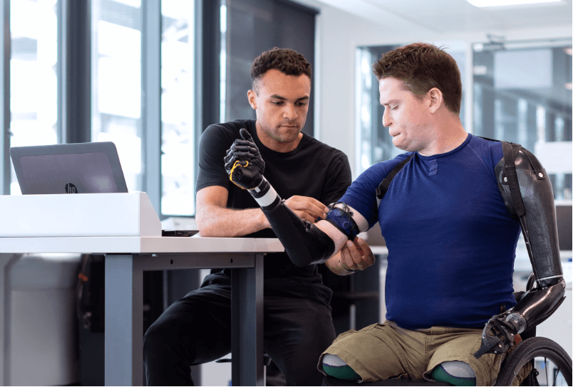 A man adjusting an armband with sensors on the upper arm of another man, who has prosthetic arms and sits in a wheelchair